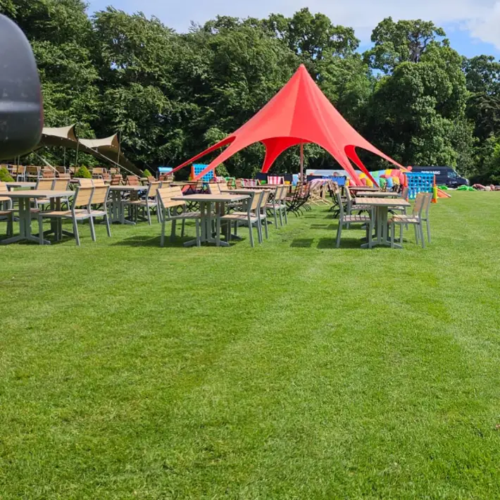Rows of wooden tables and chairs on a grassy lawn under a bright red canopy tent, with trees and event props in the background at a summer corporate gathering.