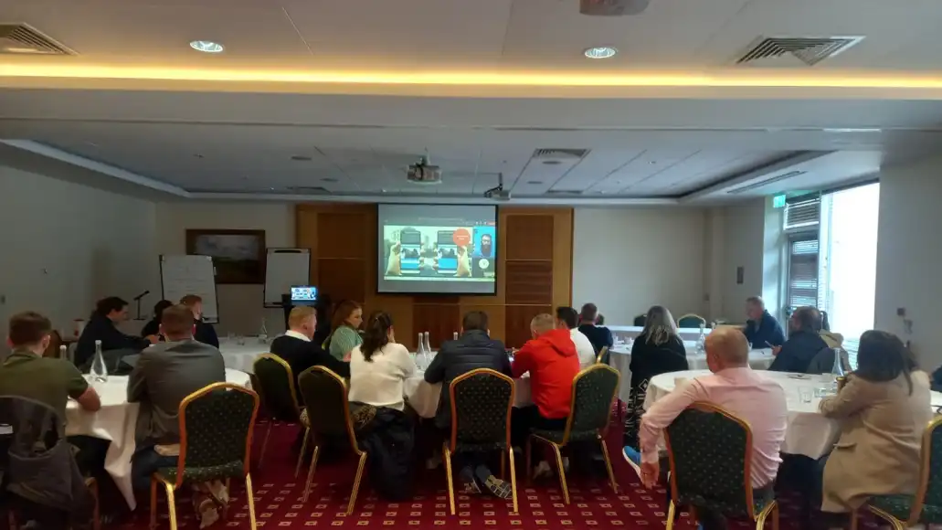 Large group of in-person employees interacting with a remote facilitator via a screen during a hybrid team event.