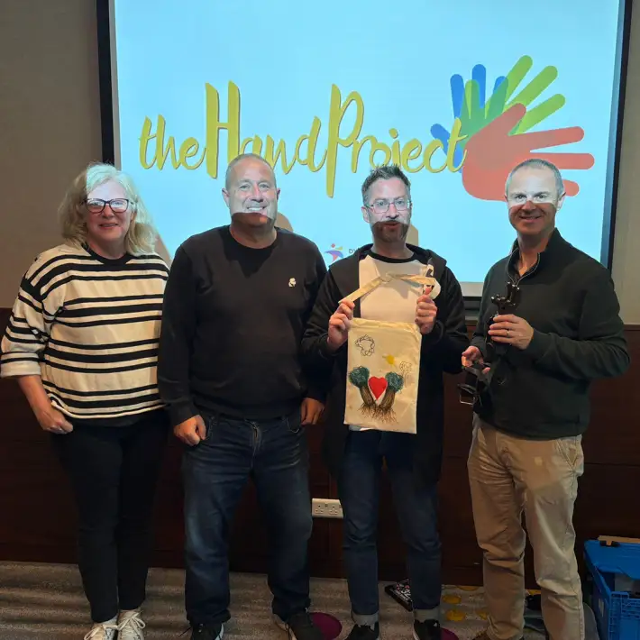 Employees taking part in The Hand Project CSR workshop, holding prosthetic hands and decorated donation bags at a corporate team building event in Ireland.