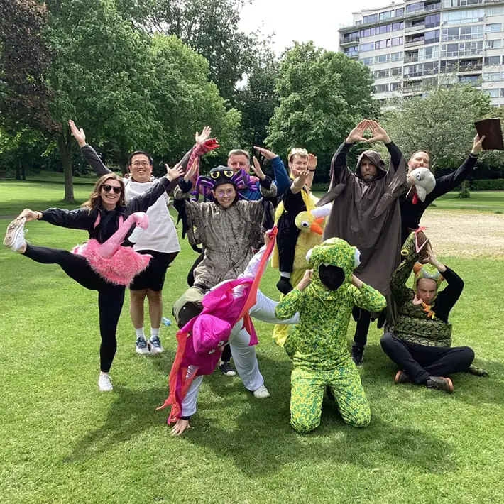 A lively group in colorful animal costumes and explorer outfits, enthusiastically posing in a grassy outdoor setting. Their dramatic gestures and vibrant outfits bring the jungle survival theme to life.