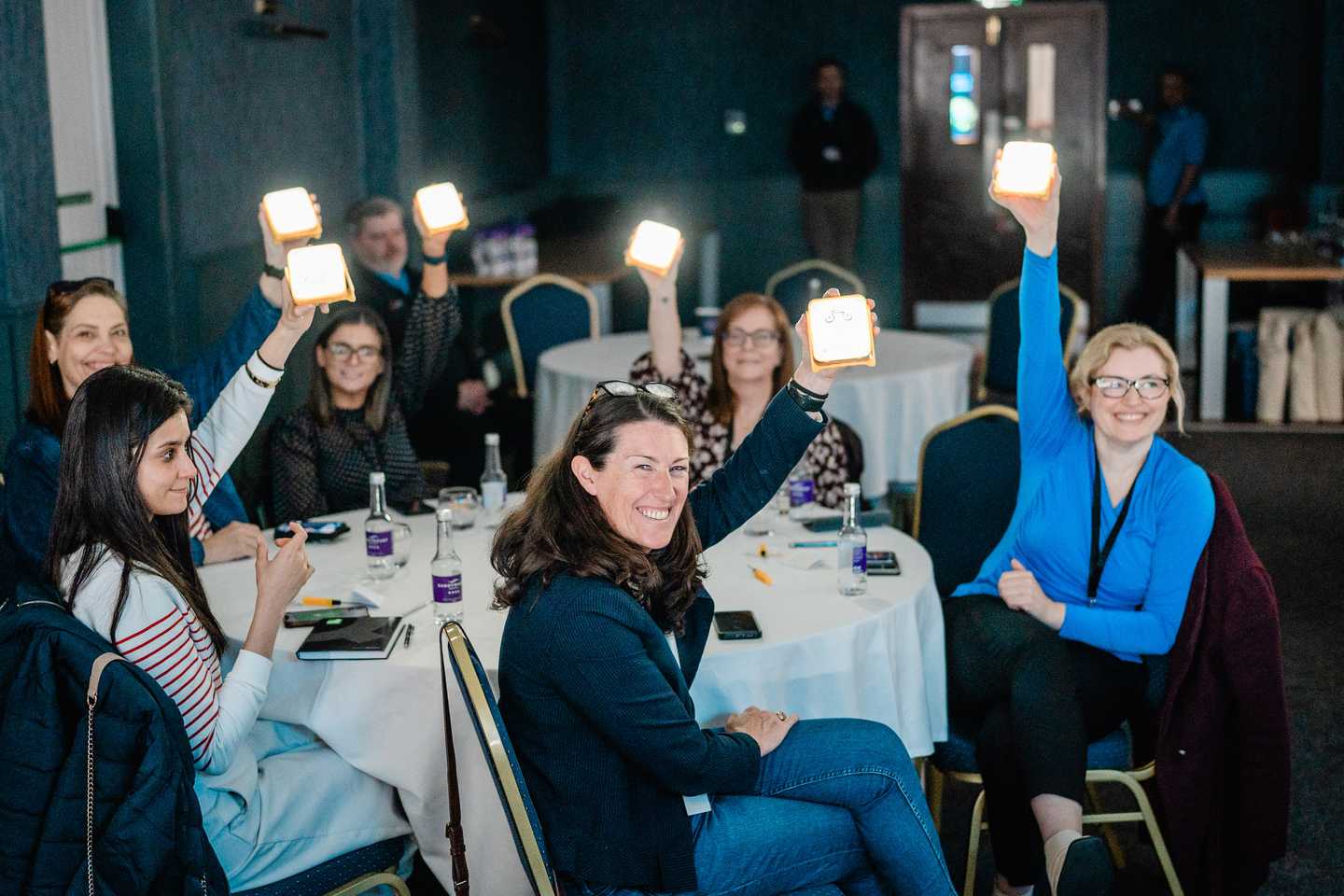 Indoor team building activity with corporate employees holding up solarbuddy lights at round tables during a facilitated team buildingworkshop in Dublin.
