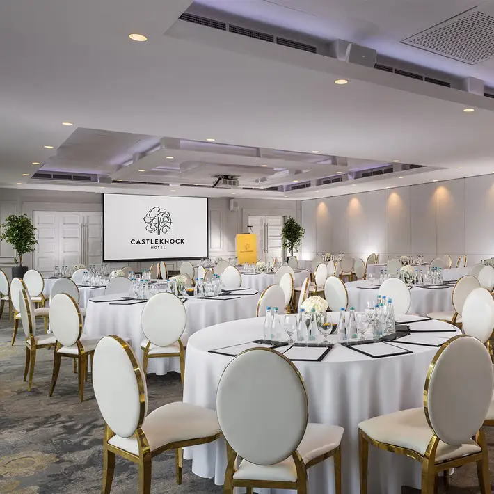 Elegant corporate event space at Castleknock Hotel, Dublin, set up for a conference with round tables, gold-accented chairs, and a large presentation screen—ideal for business meetings, conferences, and gala dinners. Event planning by Dynamic Events Ireland.