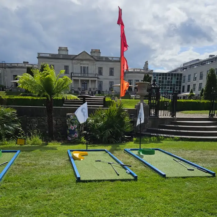 Three mini golf putting greens with clubs and obstacles arranged on a lawn, with a historic building and gardens in the background at a corporate event.