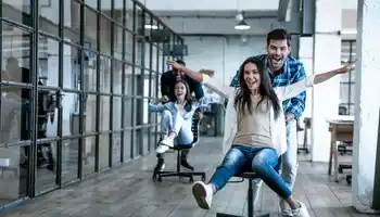Team members laughing and racing through an office on swivel chairs during a fun indoor ice breaker activity.