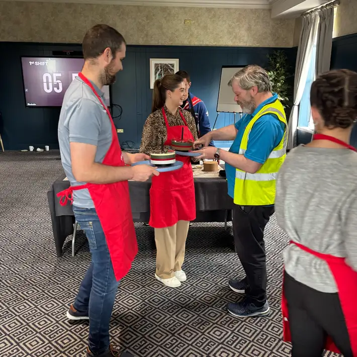 Corporate team in Ireland taking part in The Cake Factory indoor team building activity, wearing red aprons and presenting finished cakes during a quality control check.