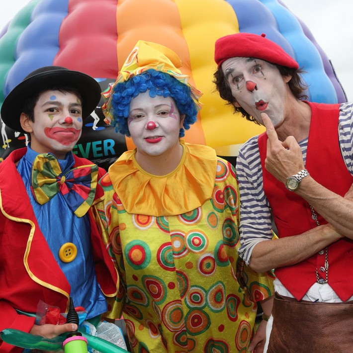 Three colourful clowns entertaining at a corporate family fun day with inflatable games in the background