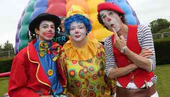 Three colourful clowns entertaining at a corporate family fun day with inflatable games in the background