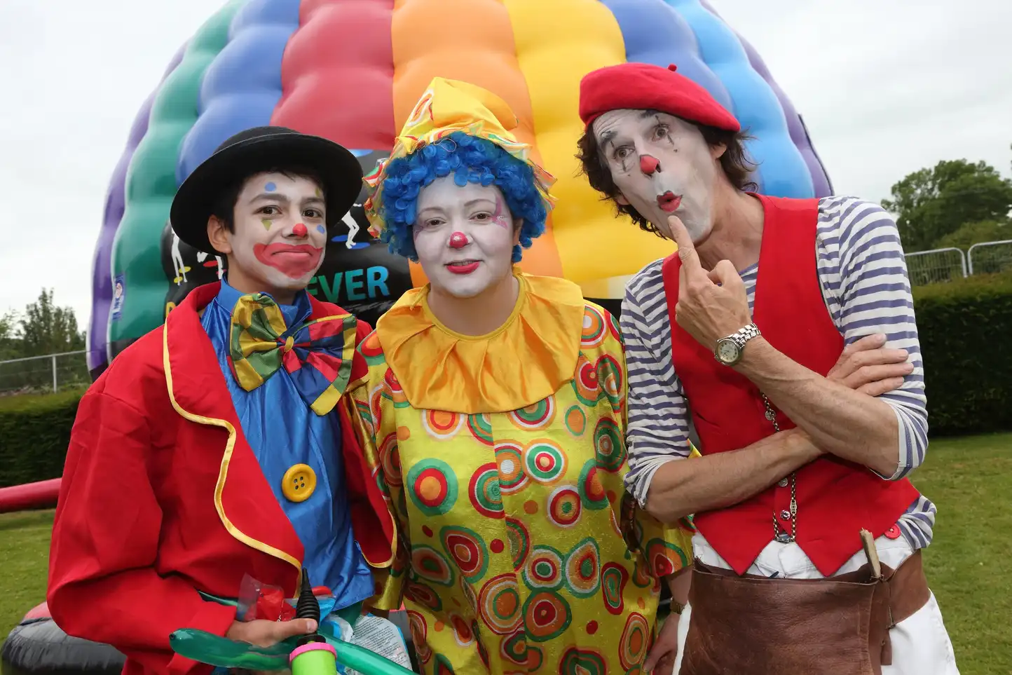 Three colourful clowns entertaining at a corporate family fun day with inflatable games in the background