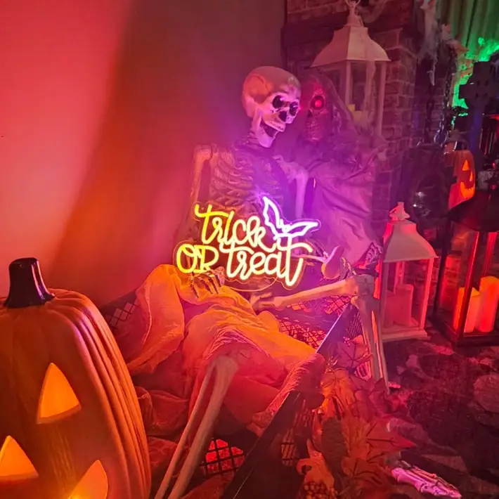 Trick or Treat neon sign sitting on a skeletons laps with picket fencing behind 