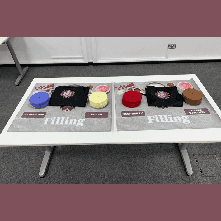 Close-up of a production station showing baking trays, recipe cards, and time-tracking sheets used during the simulation.