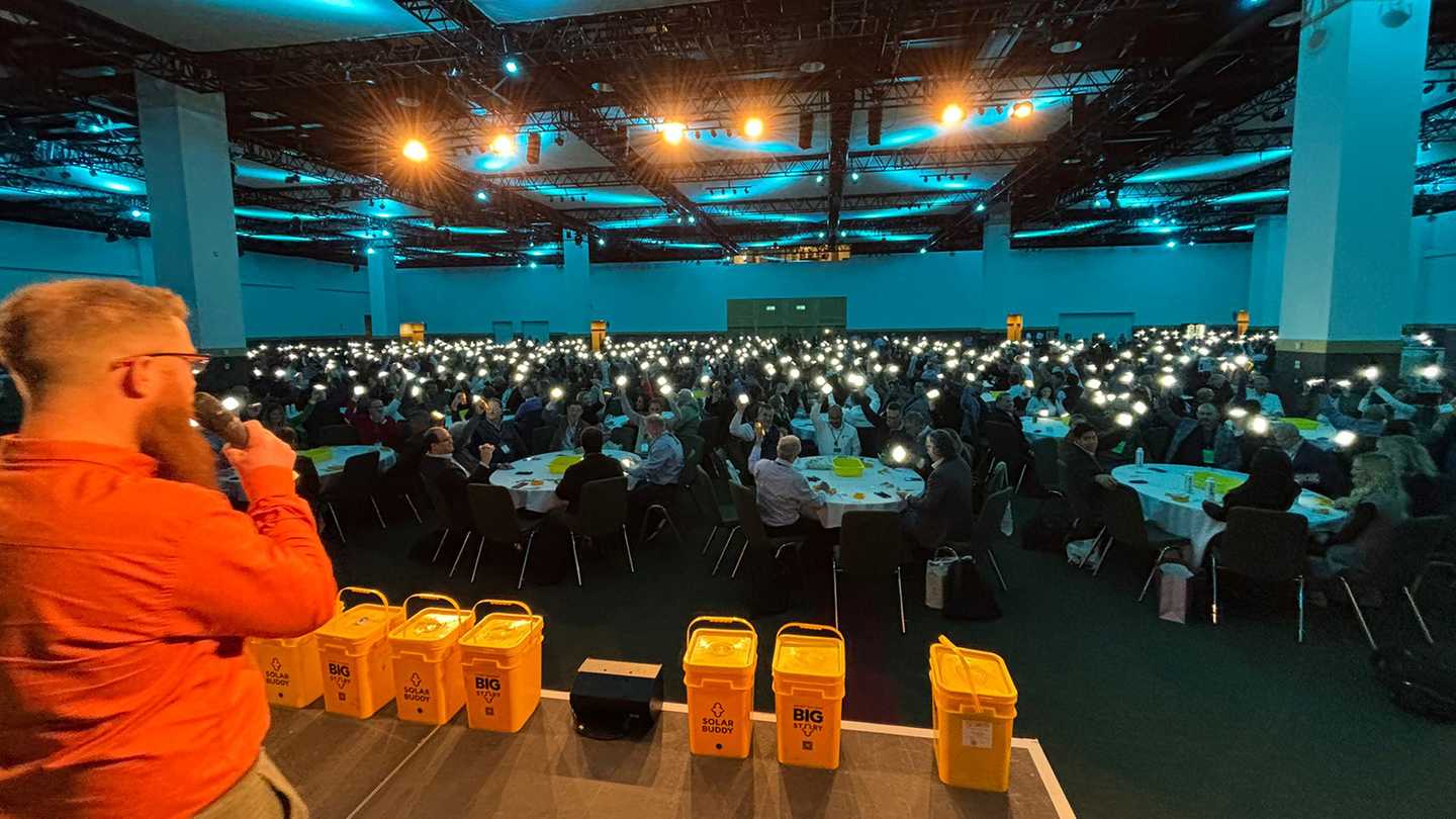 A Dynamic Events ireland host addresses a room filled with 750 Hexagon sales professionals at the Convention Centre Dublin, each holding up a glowing SolarBuddy light during the event finale. Yellow SolarBuddy collection bins are visible on stage, with the room lit by a soft blue ambience, capturing a powerful moment of unity and impact.