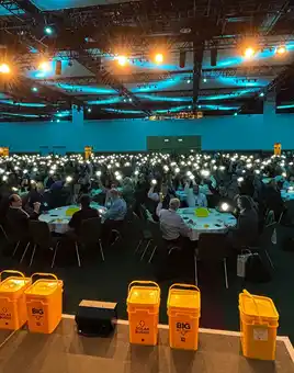 A Dynamic Events ireland host addresses a room filled with 750 Hexagon sales professionals at the Convention Centre Dublin, each holding up a glowing SolarBuddy light during the event finale. Yellow SolarBuddy collection bins are visible on stage, with the room lit by a soft blue ambience, capturing a powerful moment of unity and impact.