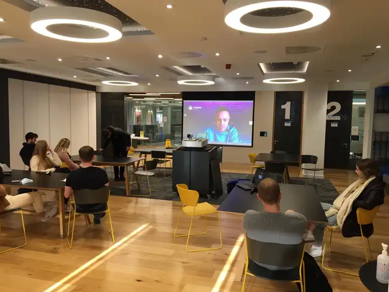 Corporate team seated in a modern meeting room receiving a remote-hosted briefing displayed on a large screen