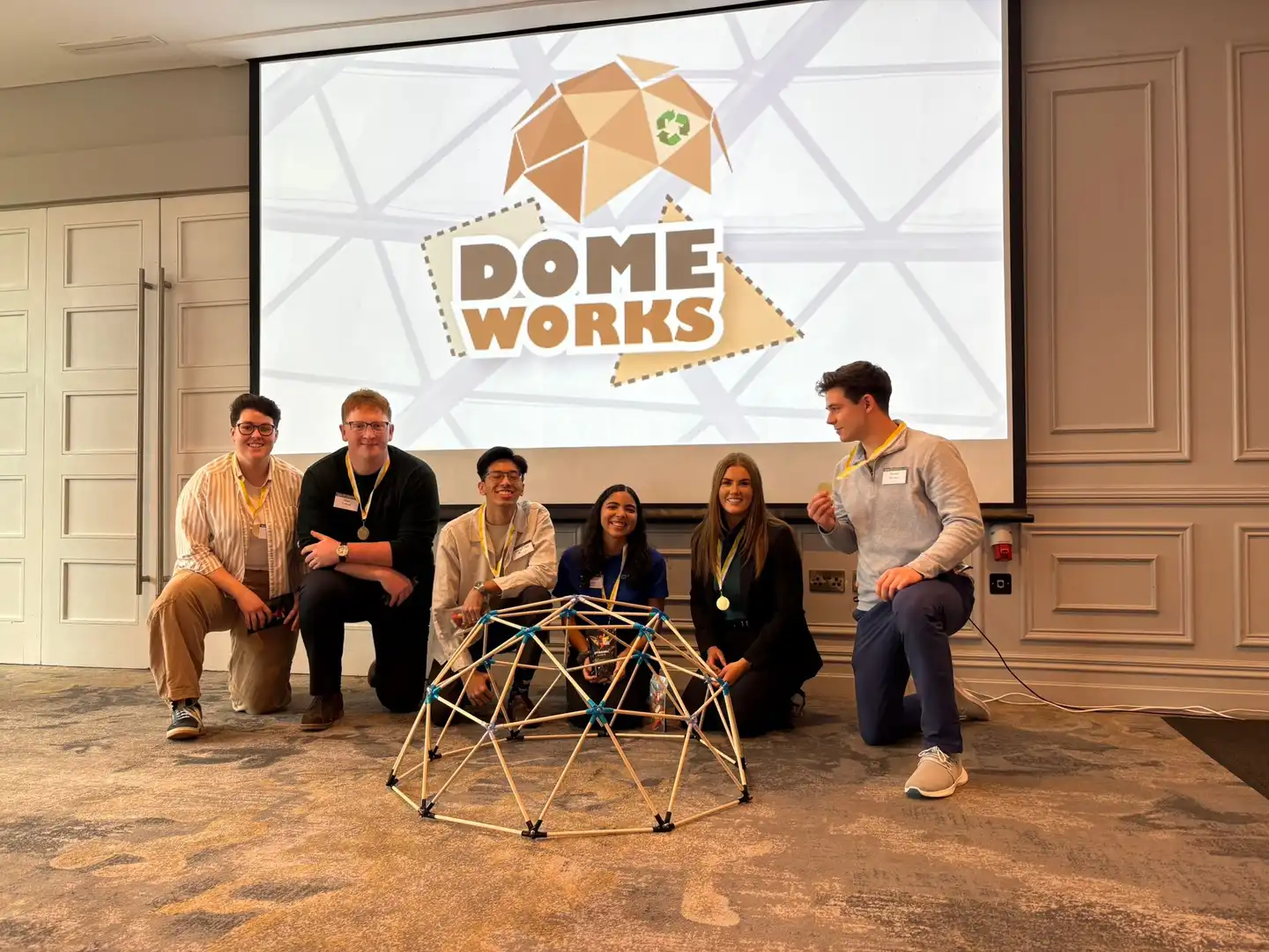 Group of graduates with the fastest time building a domeworks posing in front of the structure with medals and a Domeworks team training backdrop