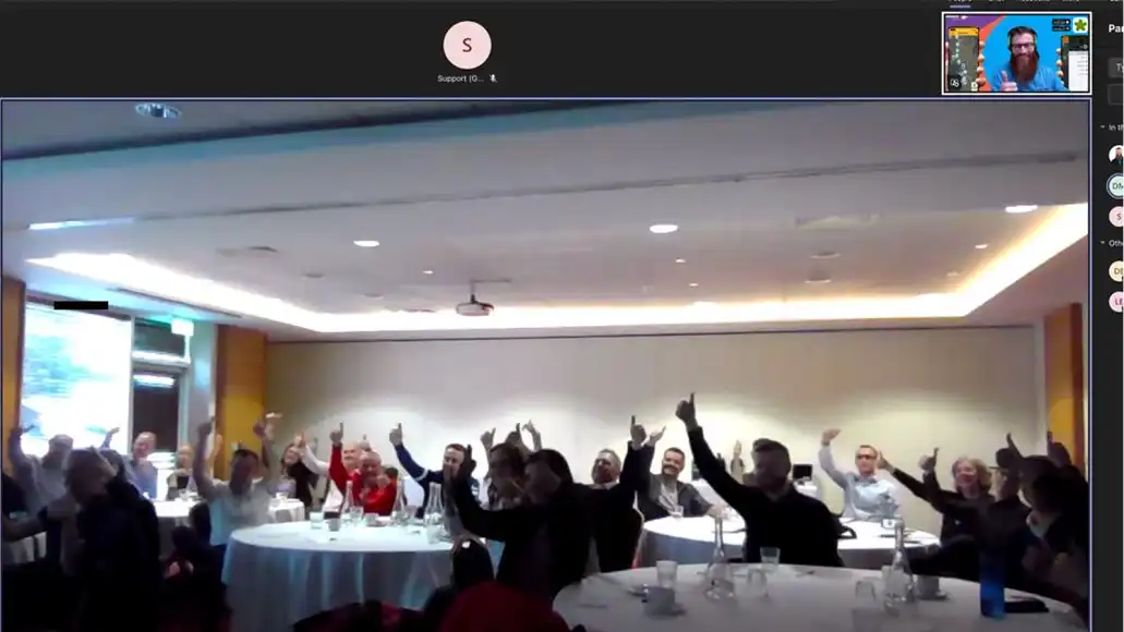 Remote host presenting an interactive hybrid team building activity while an office team collaborates at a table.