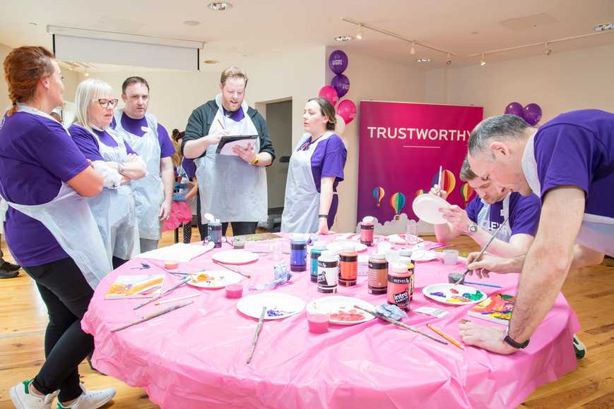 Team in purple shirts and aprons collaborating at a painting station during a group challenge