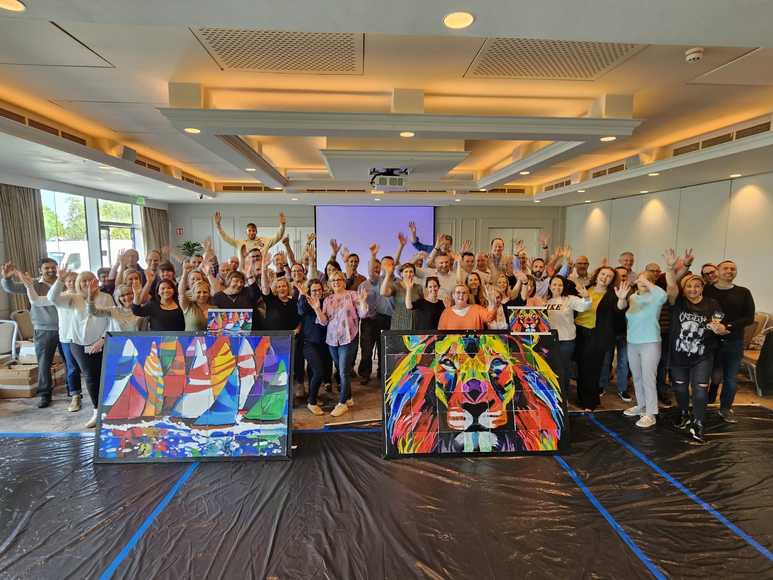 Large team celebrating in front of two collaborative murals featuring a lion and sailboats