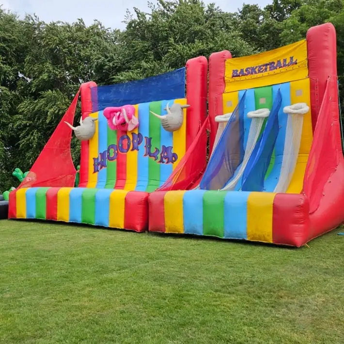 Inflatable colourful carnival-style Hoopla and Basketball game stations set up on green grass with trees in the background at a corporate summer event.