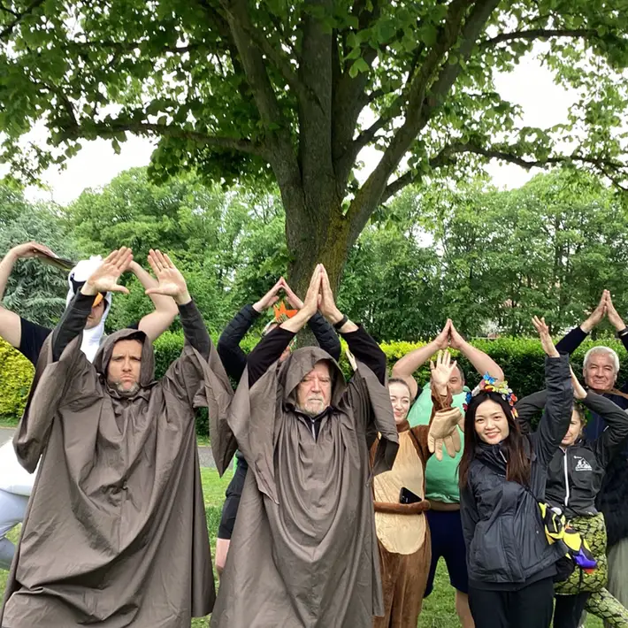 Two figures in monk-like robes stand at the center of a group, leading their teammates through a jungle survival scenario. Participants engage in synchronized poses, creating a sense of unity and adventure.