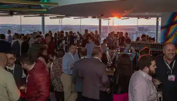 A large group of professionals enjoying a corporate networking event at sunset in Dublin, gathered around a bar serving Guinness, with a panoramic city view in the background.