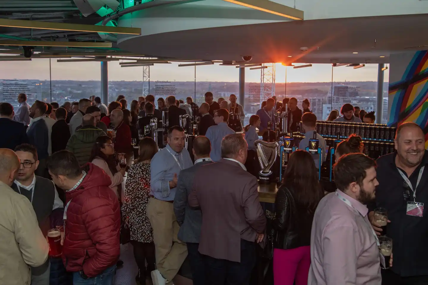 A large group of professionals enjoying a corporate networking event at sunset in Dublin, gathered around a bar serving Guinness, with a panoramic city view in the background.
