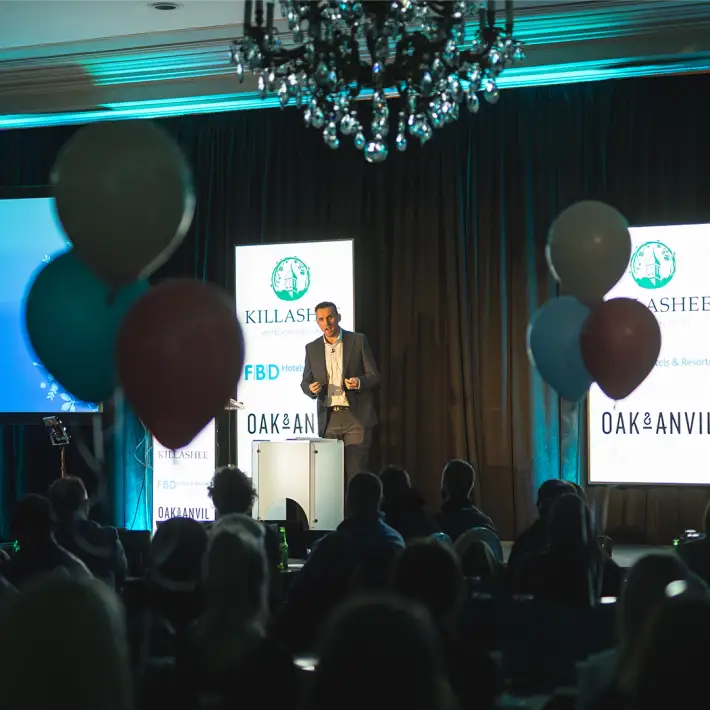 A speaker addressing employees during a company celebration focused on core values, with branded stage design and audience engagement in Ireland.