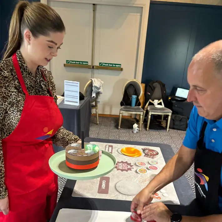 Corporate team member in red apron delivering a completed cake during The Cake Factory indoor team building event in Ireland, highlighting collaboration and strategy under time pressure.
