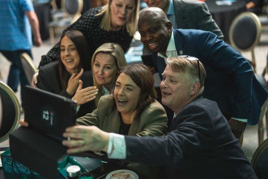 Close-up group of colleagues laughing and taking a team selfie using a tablet during an engaging workplace values event.