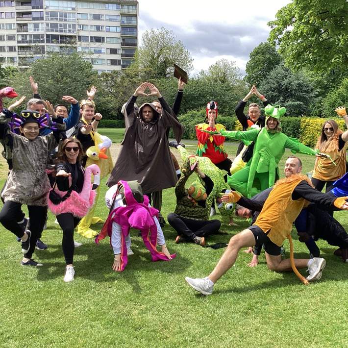 A large group of competitors dressed as jungle creatures and explorers, spread across the grass in dynamic poses, embodying the spirit of teamwork and problem-solving in their survival mission.