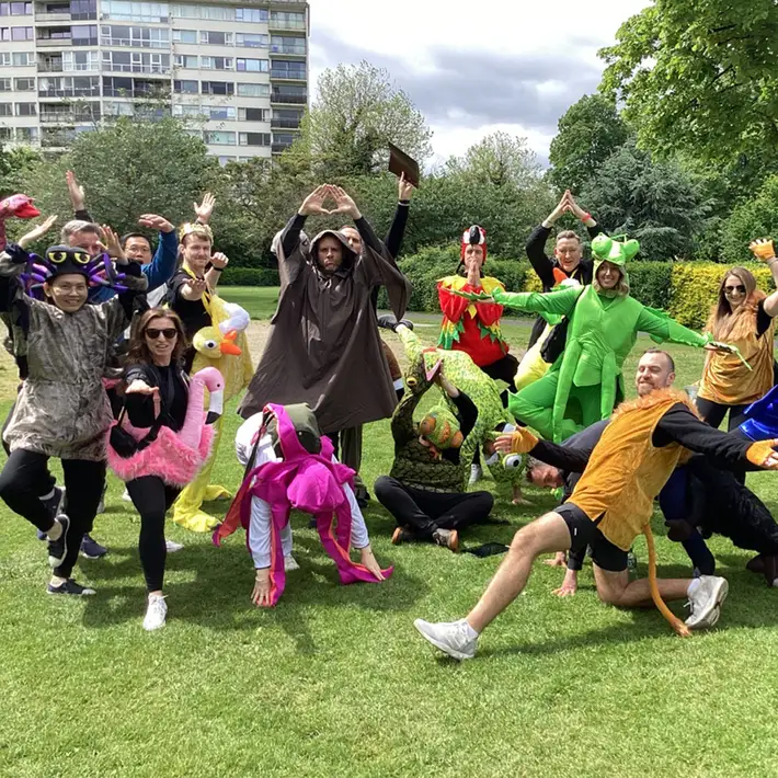 A large group of competitors dressed as jungle creatures and explorers, spread across the grass in dynamic poses, embodying the spirit of teamwork and problem-solving in their survival mission.