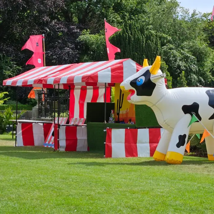 Inflatable black-and-white cow next to red and white striped carnival stalls with bunting and flags, outdoors on a sunny lawn at a corporate summer party.