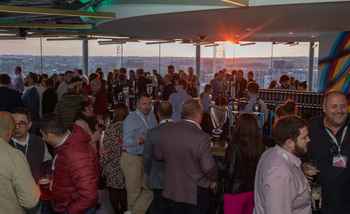A large group of professionals enjoying a corporate networking event at sunset in Dublin, gathered around a bar serving Guinness, with a panoramic city view in the background.