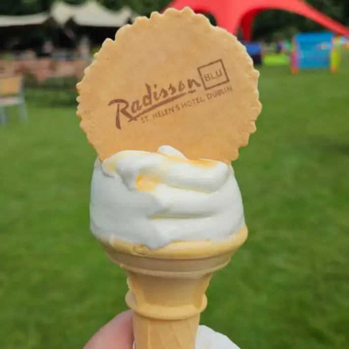Close-up of a vanilla soft-serve ice cream cone topped with a branded wafer from Radisson Blu St. Helen’s Hotel Dublin, with event lawn and tents blurred in the background.