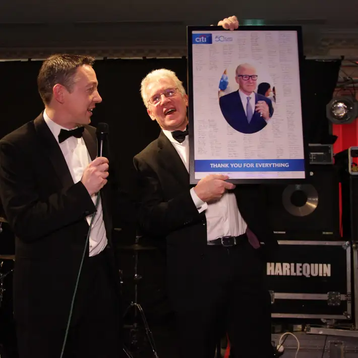 Two speakers on stage during a corporate anniversary celebration, presenting a framed tribute at a formal company event in Ireland.