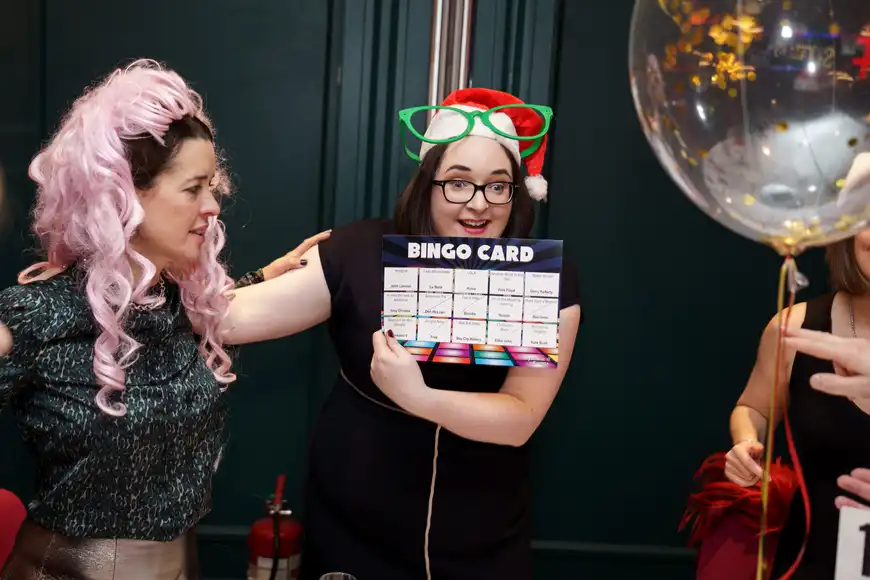 Corporate team laughing and playing Disco Bingo with festive props and Santa hats at a Christmas party.