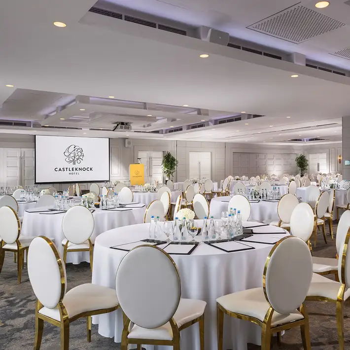 Versatile corporate event space at Castleknock Hotel, Dublin, prepared for a formal business dinner with elegantly dressed tables, floral centerpieces, and a professional presentation screen—ideal for company celebrations and award ceremonies. Delivered by Dynamic Events Ireland.