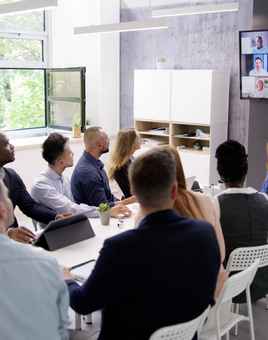 Employees sitting in a meeting room watching a Zoom call with multiple teams connected from different locations.